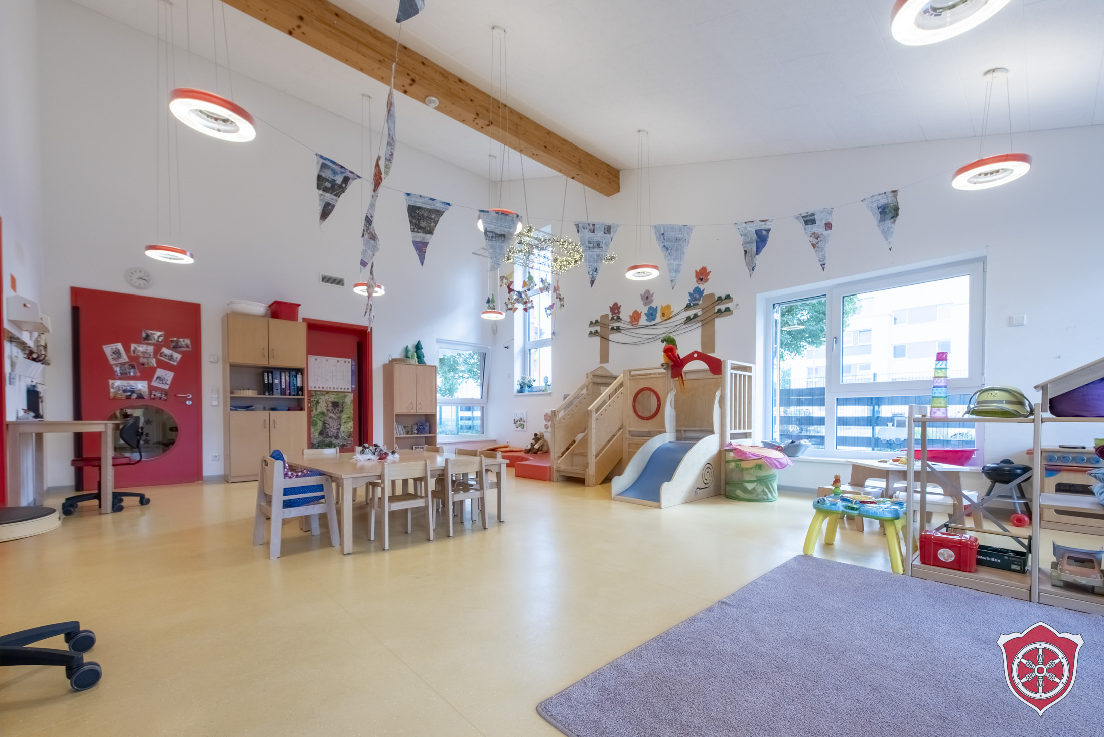 Es ist ein bunter Gruppenraum zu sehen. In ihm steht eine Tischgruppe mit Stühlen, eine Hochebene und rechts im Bild ist ein Regal mit Spielzeugen zu sehen.