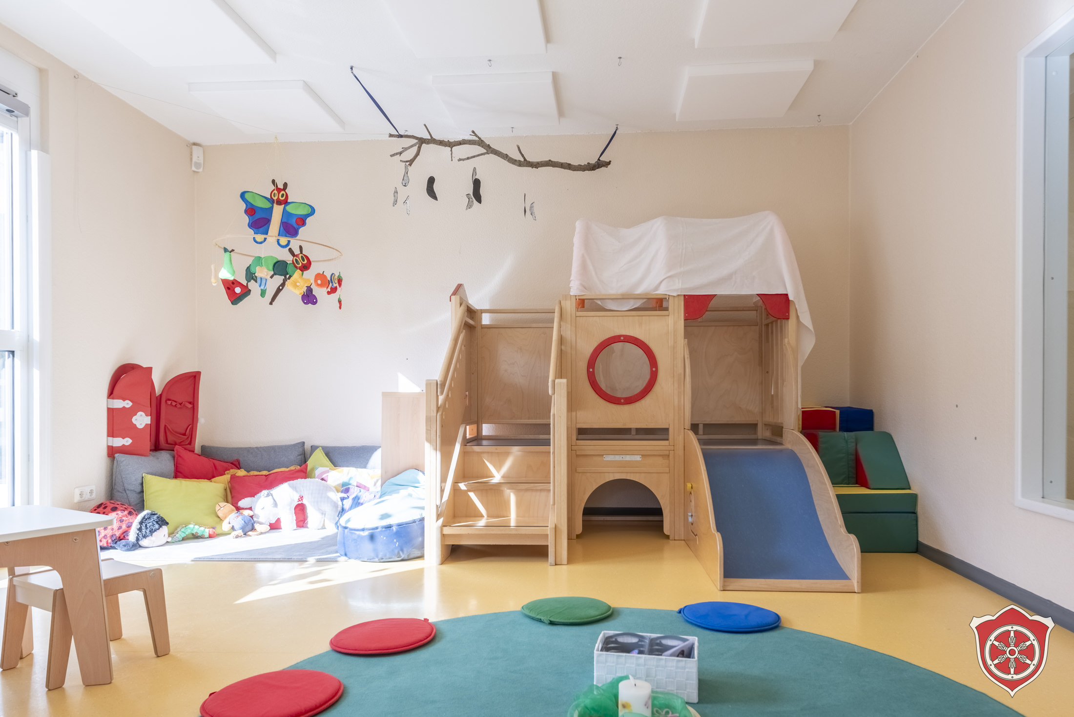 Der Gruppenraum zeigt eine Hochebene, die mit einer Rutsche ausgestattet ist. Links davon ist eine Kuschelecke. Im Vordergrund ist ein Teil eines Sitzkreises zu sehen.