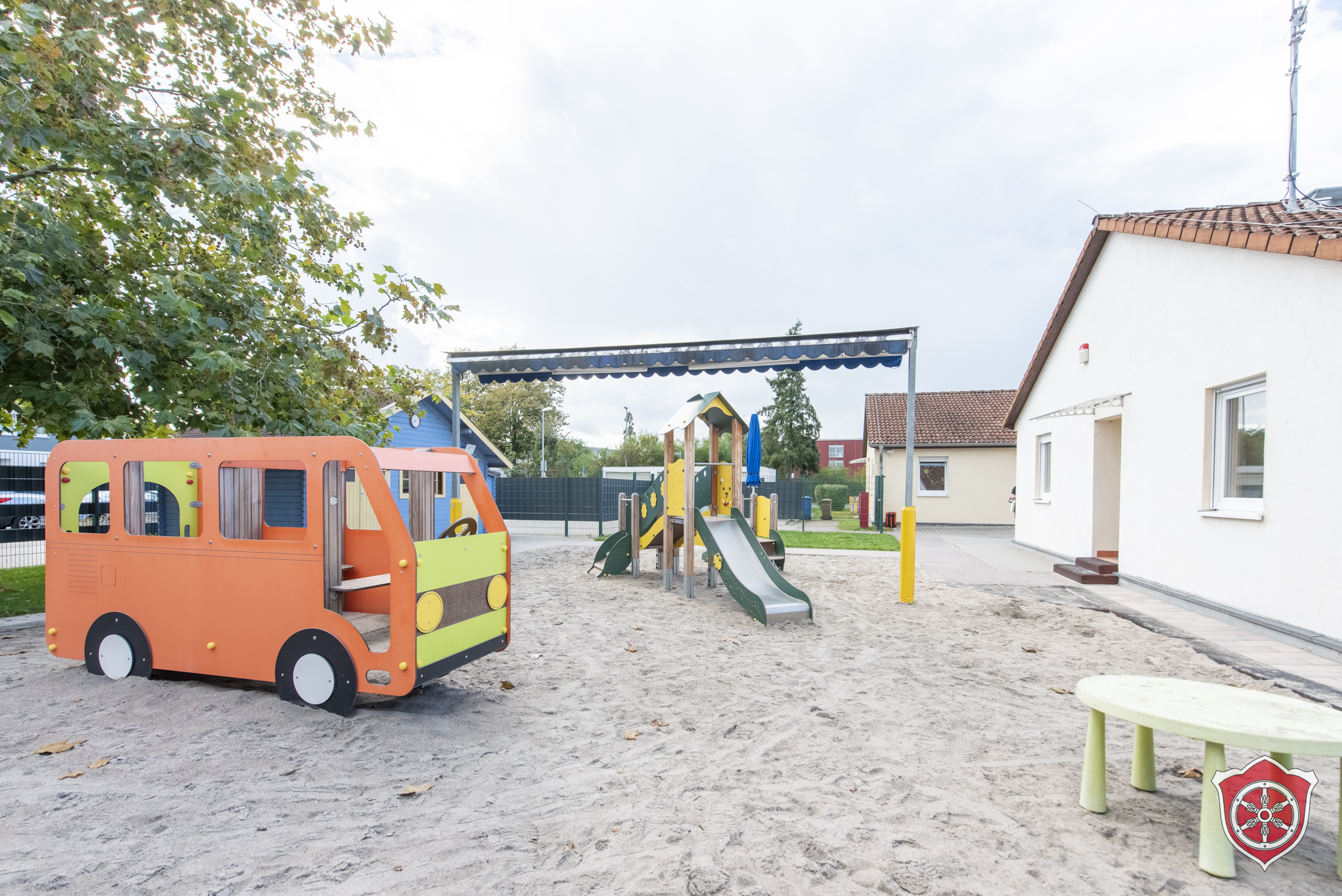 Man sieht eine Sandfläche des AUßenbereichs, Darauf steht ein Klettergerüst in Form eines kleinen Busses und eine Rutsche.