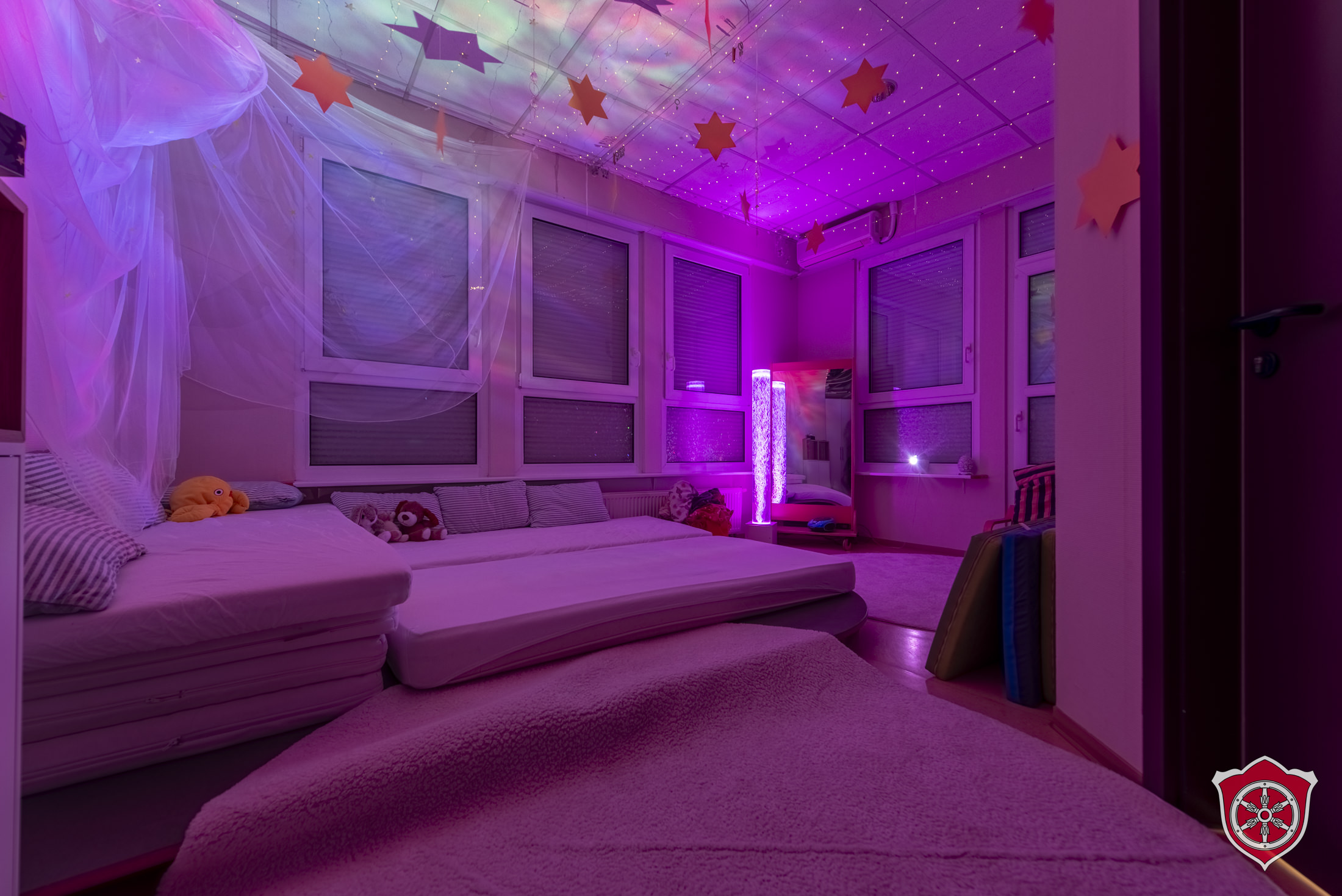 Der Snoozelraum ist ein Entspannungsraum, der abgedunkelt und in leicht lila Licht getaucht ist. Man sieht eine Kissenlandschaft und beruhigendes Licht von einer Wassersäule.