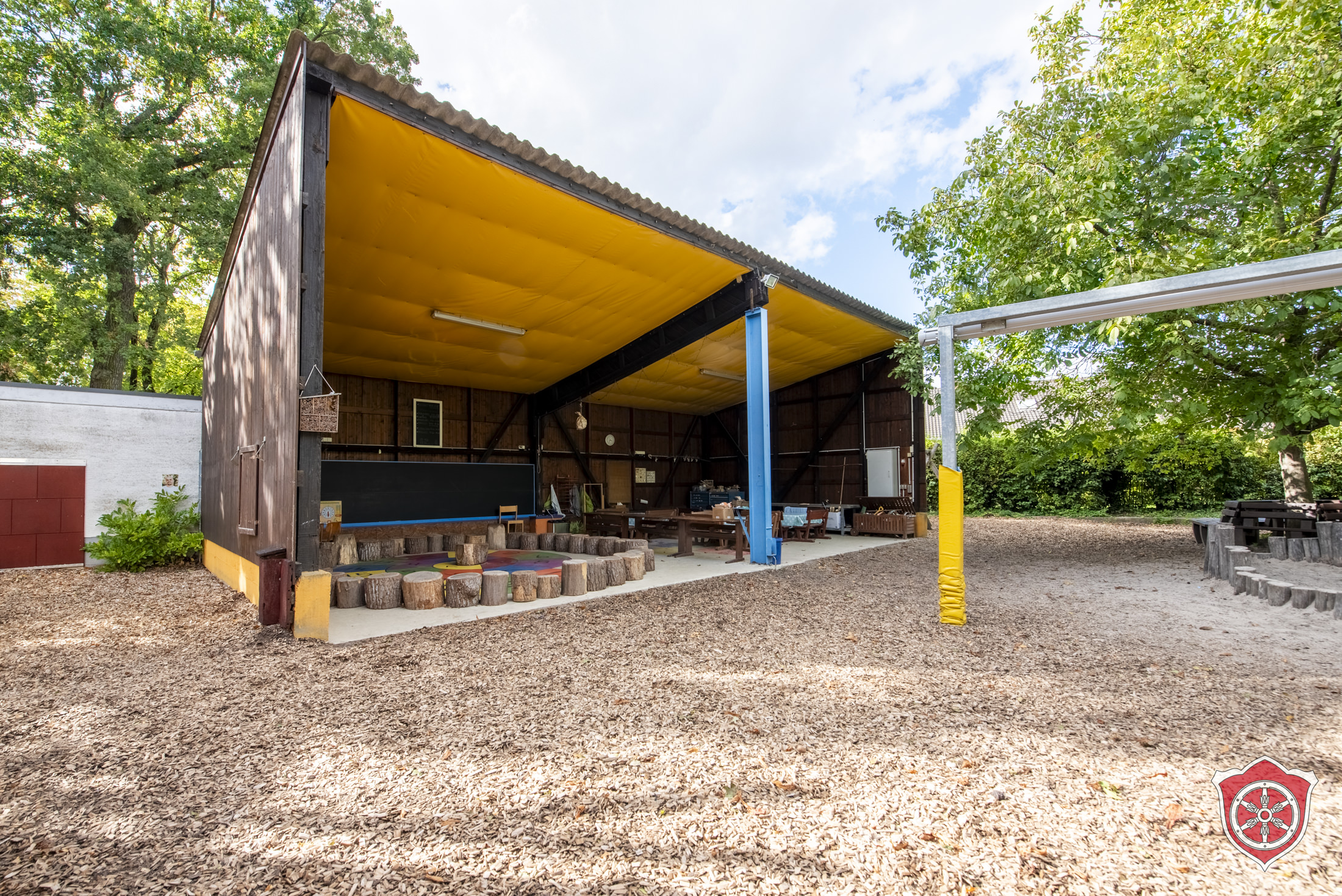 Das Bild zeigt die Basis 1. Das Carport ist zu sehen, in dem ein Stuhlkreis aus Stämmen und Tischgruppen stehen.
